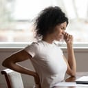 Close-up of a tired young woman at her desk, touching her lower back, experiencing pain.