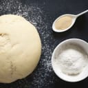 Rolled up dough, spoonful of yeast and bowlful of flour on black surface