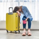 A tired woman leaning on her head on a yellow suitcase holding a red passport sleeve