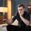 Bored guy sits on the couch in the evening in living room, holding the TV remote in his hand, switching channels, looking for a movie to watch, propping his hand under his chin, his face, unsatisfied