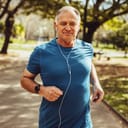 Cheerful senior man jogging in park listening to music on earphones. Senior fitness person running in park for good health.