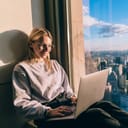 A female working on a laptop near a window of a skyscraper