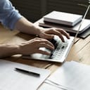 close-up of a man at a wooden desk typing on a laptop, working on documents that are next to the laptop on a desk.