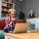 worried middle-aged woman sitting on the sofa in her living room looking at the laptop on a table