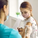 Woman doctor putting medical plaster to girl patients arm after vaccination injection against covid-19 during pandemic in clinic office. Vaccinating children against coronavirus concept.