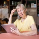 A perplexed older woman sitting at table with tablet in her hand