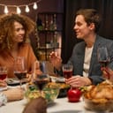 Young smiling man explaining something to his girlfriend by festive dinner while sitting among intercultural girls during communication