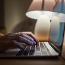 A close-up of hands typing on a laptop with a lamp in the background