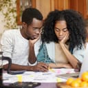 Upset young African-American couple feeling unhappy because they can't afford buying new car, facing financial problem and economic crisis, trying to solve troubles, reviewing their finances at home
