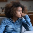 pensive teenage girl sitting on a couch at home with hand on her cheek, looking away