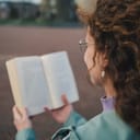 Over-the-shoulder close-up of a curly-haired woman reading a book outdoors in a spacious urban setting