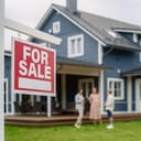 Young Couple Visiting a Potential New Home Property with Professional Real Estate Agent. Female Realtor Showing the Area to Future Homeowners. Focus on For Sale Sign.
