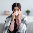 Unhappy sad young indian female in plaid suffering from fever and flu on sofa, blowing nose in napkin in living room interior. Covid-19 lockdown, treatment of illness, cold and runny, copy space
