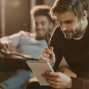 handsome young man writing lyrics in notepad while his friend lying on floor with guitar