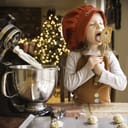A little girl in the kitchen making holiday cookies, licking the dough from a spatula.