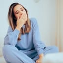 A young woman sitting on a bed holding her hand over her mouth yawning