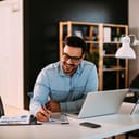 Smiling man holding a pen over some graph while working on laptop at table