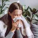 Close up of sick woman sitting on sofa freezing blowing running nose got fever caught cold sneezing in tissue, ill brunette girl covered with blanket, having influenza symptoms coughing at home