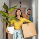 Young couple carrying cardboard boxes and a plant while moving into their new home