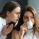 A closeup of a woman whispering something to her coworker who is drinking coffee