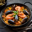 Paella in black pan with rice, shrimps, mussels, squid and meat, bowl with olives and vintage cutlery. Seafood paella, traditional spanish dish. Paella on rustic black wooden table. Selective focus