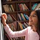 smiling female primary school student holding chalk and writing on a blackboard with bookshelf behind her