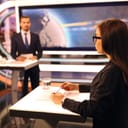 Woman in glasses and man in suit standing in studio, making a TV news show. A glass of water and paper leaf on the table. Big screen on the background.