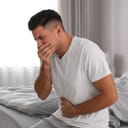 A man sitting on a bed feeling nauseous holding one hand on his stomach and the other on his mouth