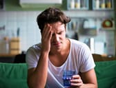 Man sitting on a couch with a glass of water in his hand holding his head with a painful expression on his face