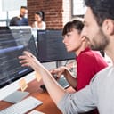 A man and woman sitting at desk looking at a monitor displaying programming code