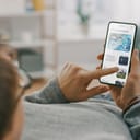 Young Man at Home is Lying on a Coach and Using Smartphone for Scrolling and Reading News about Technological Breakthroughs. He's Sitting On a Couch in His Cozy Living Room. Over the Shoulder Shot