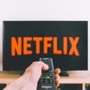 A person holding a remote control aimed at a TV screen displaying the Netflix interface.