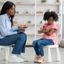 Small girl sitting on a chair with a dismissive attitude with her hands crossed next to a woman also sitting on a chair with an inquiring look on her face