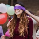 Close-up of a young girl with a blindfold at a surprise birthday party, with balloons in the background.