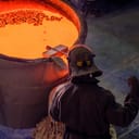 top view of a vat containing red-hot molten metal, with a worker examining its consistency before casting