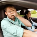 Young woman driving a car and her male passenger who covering his face with his hand in fright.