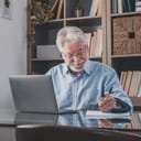 An older man sitting at desk behind a laptop taking down notes on a pad