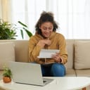 Joyful teen girl reading a university acceptance letter to her dream school in her living room, with a laptop in front of her.