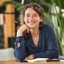 Portrait of smiling woman wearing spectacles while sitting at desk. Business woman taking notes in diary and looking at camera. University girl with eyeglasses sitting on table at library