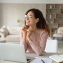 Smiling businesswoman wearing glasses daydreaming at her desk with a laptop at home office, thinking about future possibilities