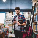 Stylish young man in reading a book's synopsis on his mobile phone while spending time in a bookstore.