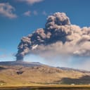 Panoramic view of a volcano erupting with thick, black smoke coming out of it high into the air
