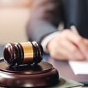 A close-up of a judge's wooden gavel on a sounding block, with a man's hand blurred in the background writing something.