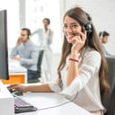 A young smiling customer support call operator speaking using a headset while seated at a computer desk