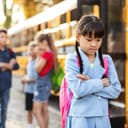 Little Asian schoolgirl looking sad and angry near a school bus while classmates chat in the background