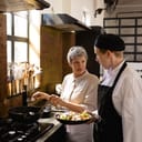 older female chef talking with a younger male colleague who is holding a plate of chopped vegetables in a restaurant kitchen