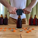 Craft beer brewing at home, man closes brown glass beer bottles with plastic capper on wooden table with orange crown caps. Horizontal image