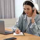 young woman with headphones taking notes at a table while listening to the lecture