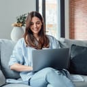 A concentrated millennial girl sitting on a couch working on the laptop, browsing internet and smiling.