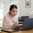 Girl with earphones at table taking down notes in front of laptop